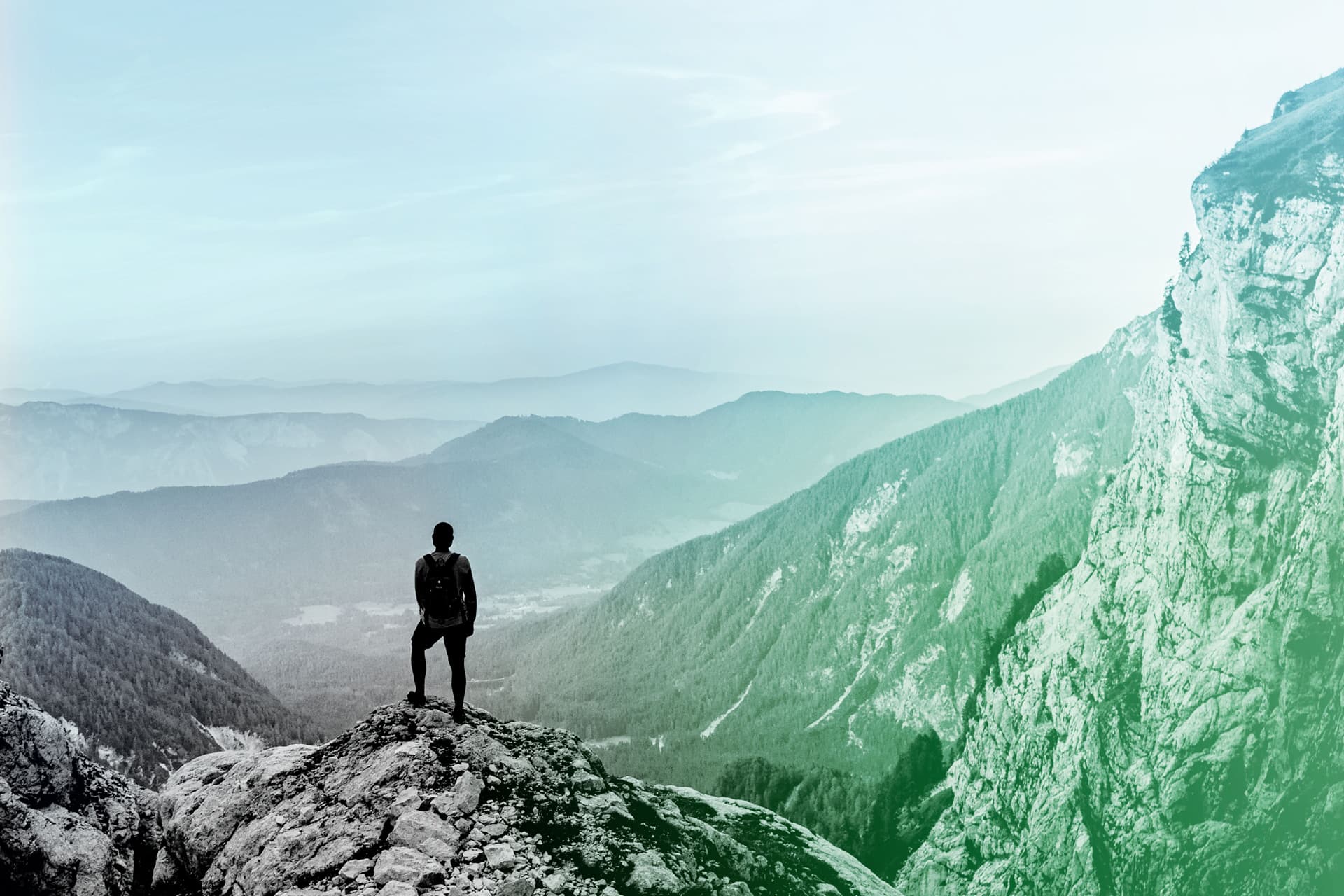 Person steht auf Berggipfel mit Blick über nebelbedecktes Tal und Berglandschaft.